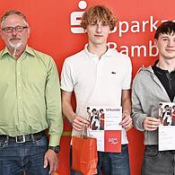 Vorstandsvorsitzender Stephan Kirchner (links) und Wertpapierbeauftragter David Förtsch (rechts) gratulieren Samuel Ziegelhöfer und Leon Motschenbacher vom Siegerteam „Gejagt vom Finanzamt“ vom Clavius-Gymnasium, zusammen mit ihrem Lehrer Jürgen S...