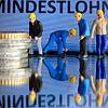 Miniaturfiguren stehen vor einem Display mit der Anzeige „Mindestlohn“.